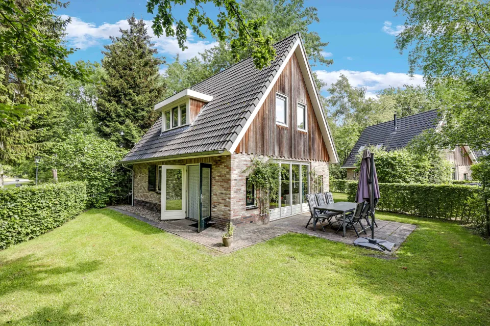 Vrijstaande vakantievilla met houten gevel, terras met tuinmeubelen en parasol in een groene tuin.